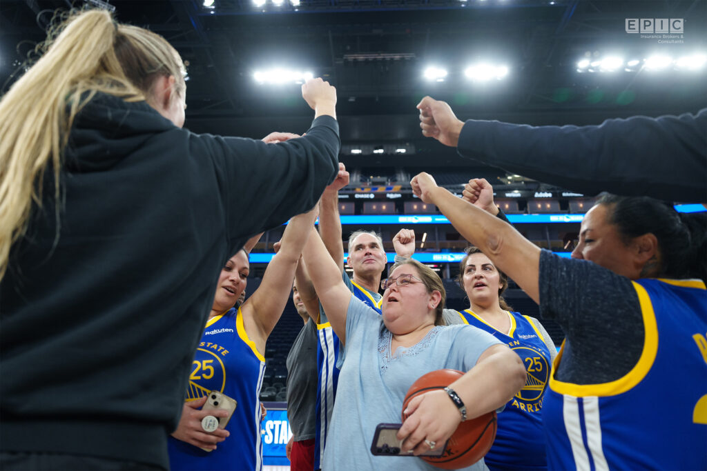 Team members in Golden State Warriors jerseys celebrate with a group fist bump in a basketball arena.